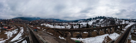 Viaduct railway bridge in the Carpathians of Ukraine, a view of the bridge from a height, a winter panorama of a village in the mountains.の写真素材