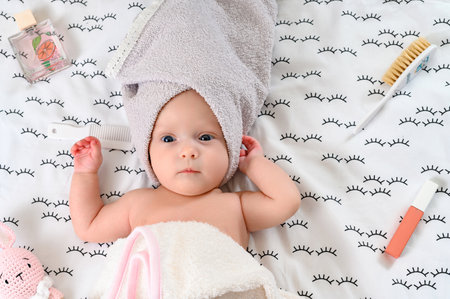 Ivano-Frankivsk, Ukraine May 8, 2022: A cute little girl wrapped in towels lies between toys and holds a comb in her hands, baby procedures after bathing.のeditorial素材