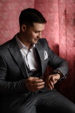 Ivano-Frankivsk, Ukraine July 31, 2022: A man with a glass of cognac in a classic suit sits by the window, the morning of the groom before the wedding.のeditorial素材