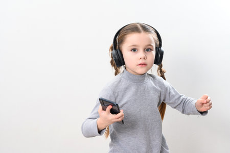 Happy little girl with wireless headphones and phone, portrait of girl with pigtails on white background with headphones and smartphone, photo with copy space.の写真素材