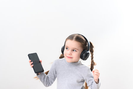 Happy little girl with wireless headphones and phone, portrait of girl with pigtails on white...