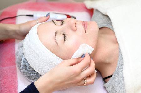 A woman cosmetologist washes a client with cotton wool and water, the client is wearing a disposable cap and is lying on the couch.の写真素材