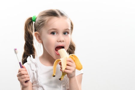A little girl eats a banana and holds a toothbrush in her hands, studio photo on a white background, a banana helps against caries, an idea for dentistry.の写真素材