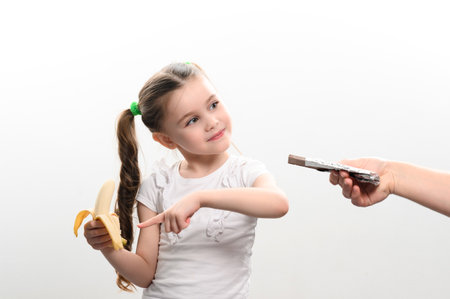 little girl shows with finger that she doesn't want chocolate and chooses banana, white background and copy space, healthy and unhealthy food concept.の写真素材