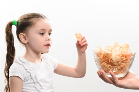 Little girl is given a big bowl of chips snacks with lard, white background portrait of a little girl eating chips, a child and unhealthy fast food.の写真素材