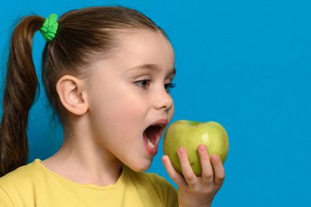 A little girl holding and biting a healthy green apple on a blue background, teeth and healthy nutrition.の写真素材