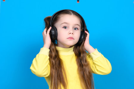 A girl on a blue background in a yellow sweater with headphones on her head, wireless headphones in a child, conceived childの写真素材