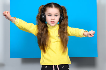 Happy little girl with wireless headphones on blue background doing dance moves to music in headphones, child with long hair.の写真素材