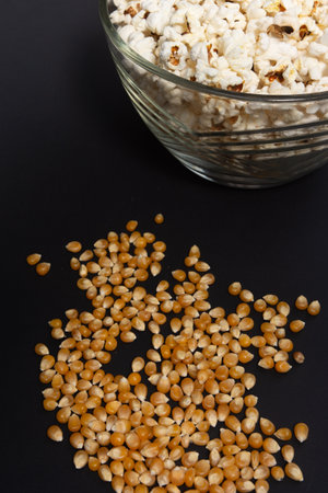 Ready-to-eat popcorn in a glass bowl and corn kernels on a black background, top view and copy...