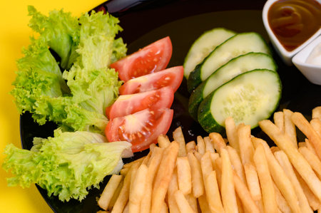 Plate of french fries with salad, tomatoes and cucumbers, sauces in white french fries plates, fast food on yellow background.の写真素材