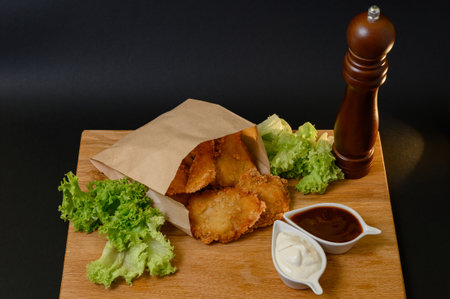 Serving of chicken nuggets on wooden board with pepper, mayonnaise and ketchup sauce, lettuce, food and black background with chicken nuggets.の写真素材