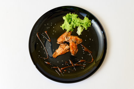 Caramelized chicken wings on a plate with salad and barbecue sauce, black plate and white background.の写真素材