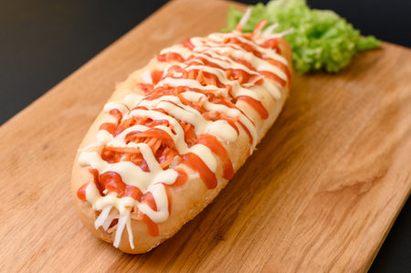 Hot dog covered with ketchup and mayonnaise on a wooden board on a black background with green salad leaves.の写真素材