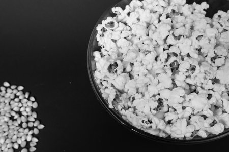 Ready-to-eat popcorn in a glass bowl and corn kernels on a black background, top view and copy space.の写真素材