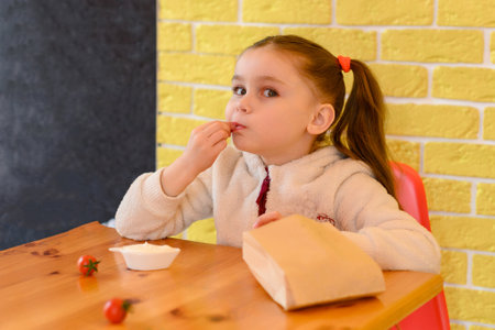 A small and beautiful girl eats french fries in a cafe, french fries in a paper bag.の写真素材