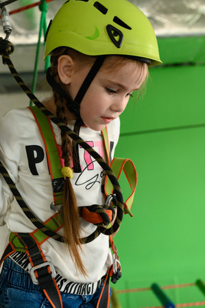 A little girl is walking on a cablewaya playroom with a cable car, a helmet and a belay is worn by a...