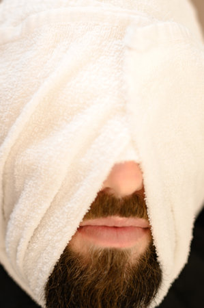 A client is lying on a chair in a barbershop with his beard wrapped in a towel. Steaming the beard before the royal shave.の写真素材