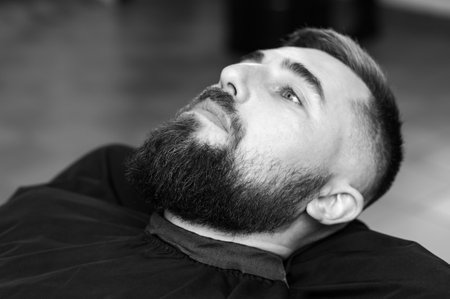 A young man with a beard in a black cape sits on a chair in a barbershop. The client is waiting for a haircut and shaving of the beard.の写真素材