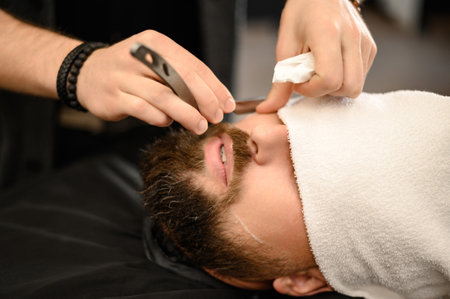 A barber shaves the cheek of a bearded customer with a dangerous razor. Shaving the contour of the beard for the correct shape.の写真素材