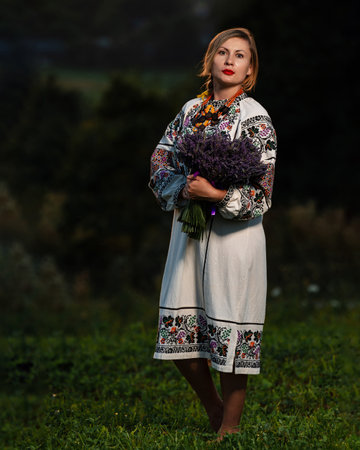 A young girl wearing a long embroidered shirt holds a lavender bouquet in her hands, photo at sunset.の写真素材