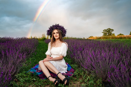 Ivano-Frankivsk, Ukraine July 28, 2023:A girl with a lavender wreath on her head sits on the grass between rows of lavender, with a rainbow in the sky in the background.のeditorial素材