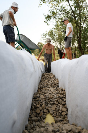 A man pours rubble crushed stone from a wheelbarrow into a trench. Drainage works for drainage of ground water around the field.の写真素材