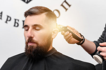 A barber cuts a man with a beard in a barber shop. Short haircut of the client with a clipper.