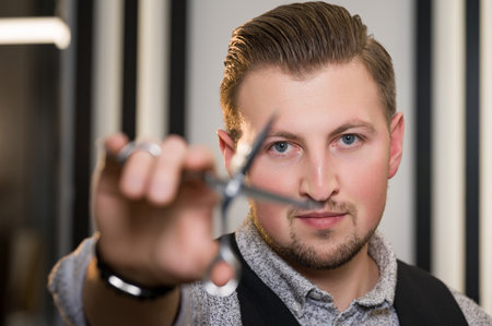 A young Caucasian barber holds scissors in his hand in front of his face. Barbershop master concept.の写真素材