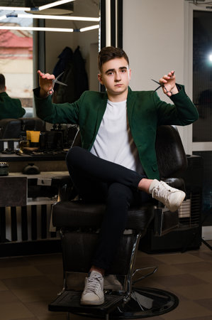 The stylist holds scissors in both hands, which are an attribute of a hairdresser. Portrait of a young barber in a jacket and white t-shirt.の写真素材