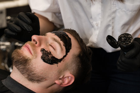 Barber applies a black moisturizing mask to a mans face while shaving. A dermatologist applies a black mask to the face of a bearded patient to clear pores and wrinkles.の写真素材