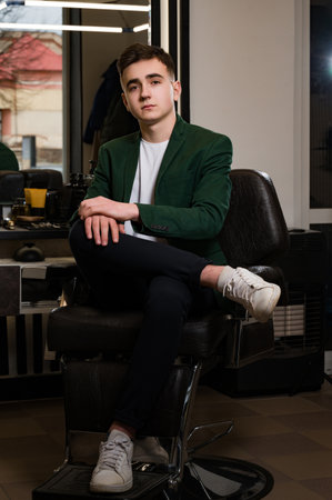The client is sitting in a barbershop chair after a haircut, satisfied with the result. Portrait of a young barber in a jacket and white t-shirt.の写真素材