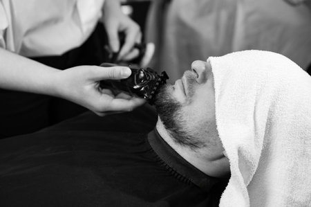 The client's head is covered with a towel during a beard trim. Shortening the length of the beard from the sides by the master for the client.の写真素材