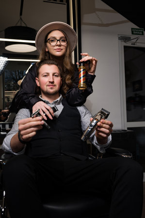 Barber boy and girl are holding tools with barbershop attributes sitting on a chair in the salon.の写真素材