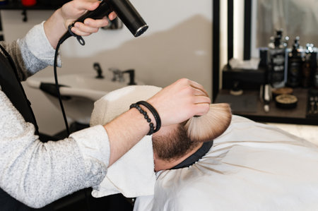 The hairdresser sweeps the hair during the beard trimming with a brush and cold drying air.の写真素材