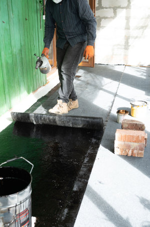 A man unrolls a hot and heated roll of bitumen, pushing with his foot, waterproofing the floor in the house.の写真素材