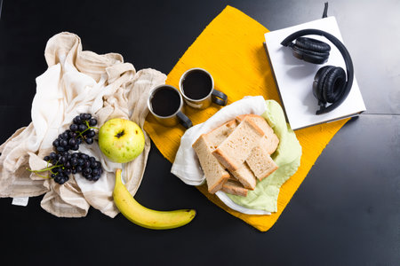 Breakfast or snack spread with fruit, bread, coffee, and headphones on a dark background.の写真素材
