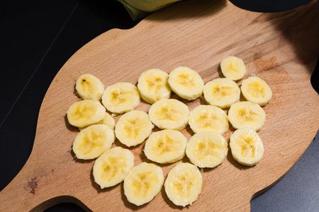 Freshly sliced banana pieces arranged on a wooden cutting board, ready for consumption or use in a recipe. The vibrant yellow color of the fruit contrasts with the natural wood.の写真素材