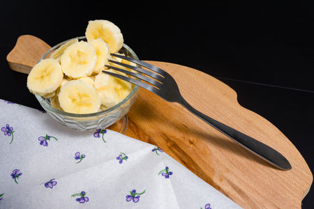 Freshly sliced banana pieces are served in a clear glass bowl. A metal fork rests on the wooden cutting board next to the bowl, ready to enjoy this simple and healthy snack.の写真素材