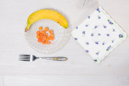 A simple table setting featuring a banana, a small portion of orange berries on a glass plate, a decorative napkin, and a fork on a white wooden table.の写真素材