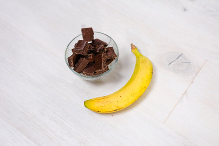 A clear glass bowl filled with broken pieces of aerated dark chocolate sits beside a single, ripe yellow banana on a light, textured wooden surface.の写真素材