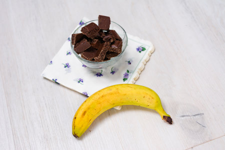 A clear glass bowl filled with broken dark chocolate pieces sits on a floral napkin. Beside it, a ripe yellow banana rests on the light-colored wooden table.の写真素材