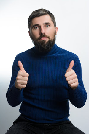 A bearded man wearing a blue turtleneck sweater and dark pants sits against a light background, giving two thumbs up with a positive expression.の写真素材