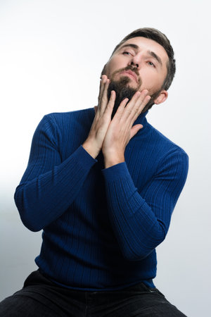 A man with a beard, wearing a blue sweater and dark pants, gently touches his facial hair against a light background.の写真素材