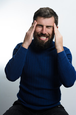 A man with a beard, wearing a blue sweater, grimaces in pain while holding his head. He appears to...