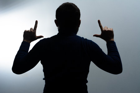 Dark silhouette of a person's back, arms raised, gesturing with their hands against a light gray backdrop. Focus on body language and shape.の写真素材