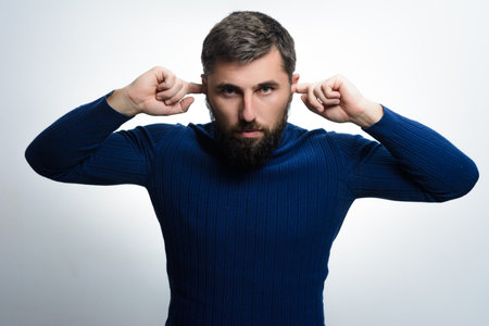 A man with a beard, wearing a blue sweater, covers his ears with his fingers, conveying a sense of frustration or a desire to block out noise.の写真素材