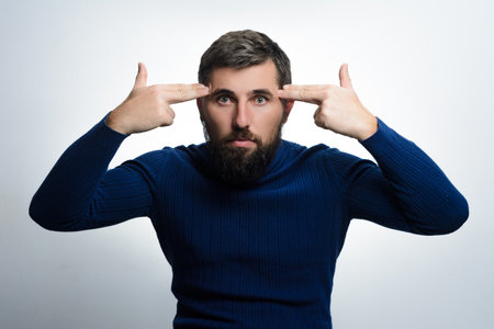 Bearded man gestures with fingers pointed at his temples against a plain background, suggesting frustration or troubled thoughts.の写真素材