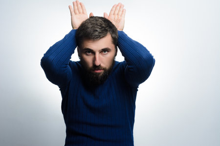 A bearded man in a blue sweater playfully gestures with his hands above his head, forming bunny ears against a light gray background.の写真素材