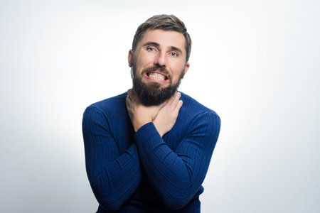 A bearded man with a pained expression clutches his throat with both hands against a light background. He appears to be choking or experiencing discomfort.の写真素材