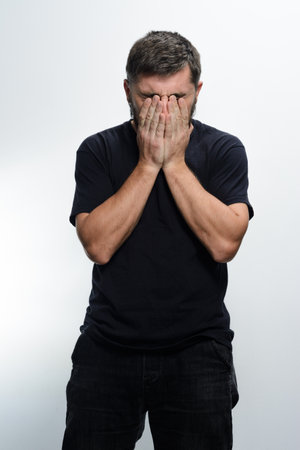 A man with dark hair and a beard stands against a light background, his face buried in his hands, conveying a sense of sadness, shame, or frustration.の写真素材